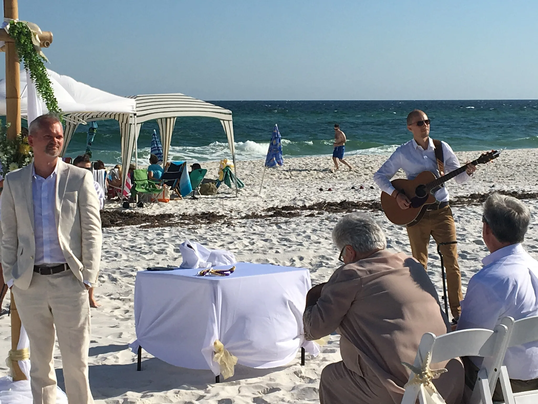 Matt Headley playing acoustic guitar during a beach wedding ceremony as the couple stands at the arch