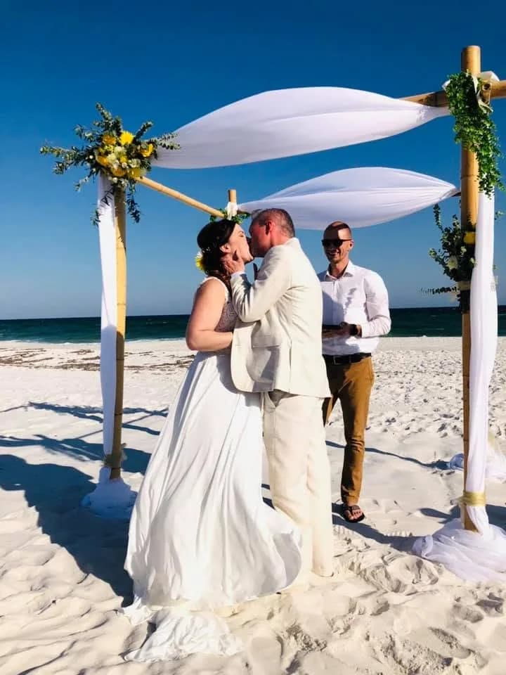 A bride and groom kiss under a bamboo arch on the beach, Matt Headley officiating beside them