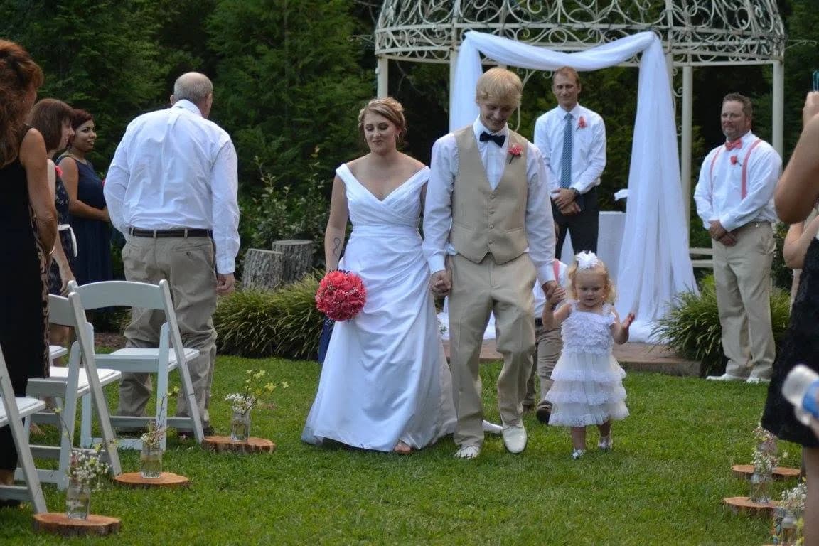 A newly married couple walk back down the aisle at an outdoor garden ceremony, Matt Headley at the gazebo behind them