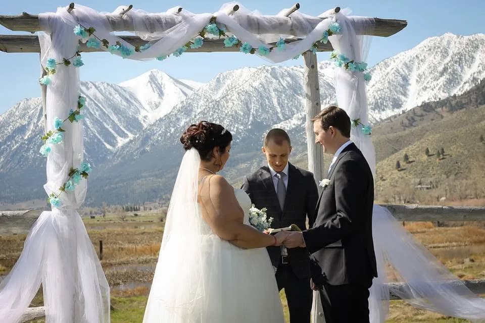Matt Headley officiating an outdoor wedding ceremony as the couple exchanges rings, snow-capped mountains behind them
